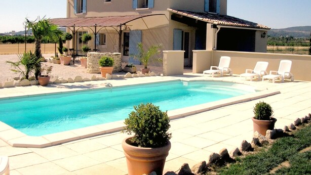 Una casa de dos pisos con piscina, mobiliario de exterior y plantas en macetas.