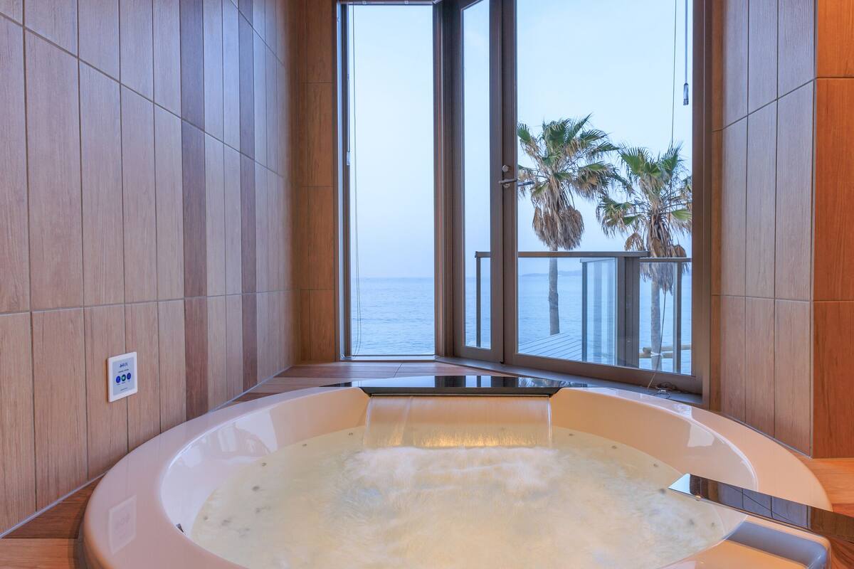 A spa area with a hot tub, wooden walls, and a view of palm trees and the sea through large windows.