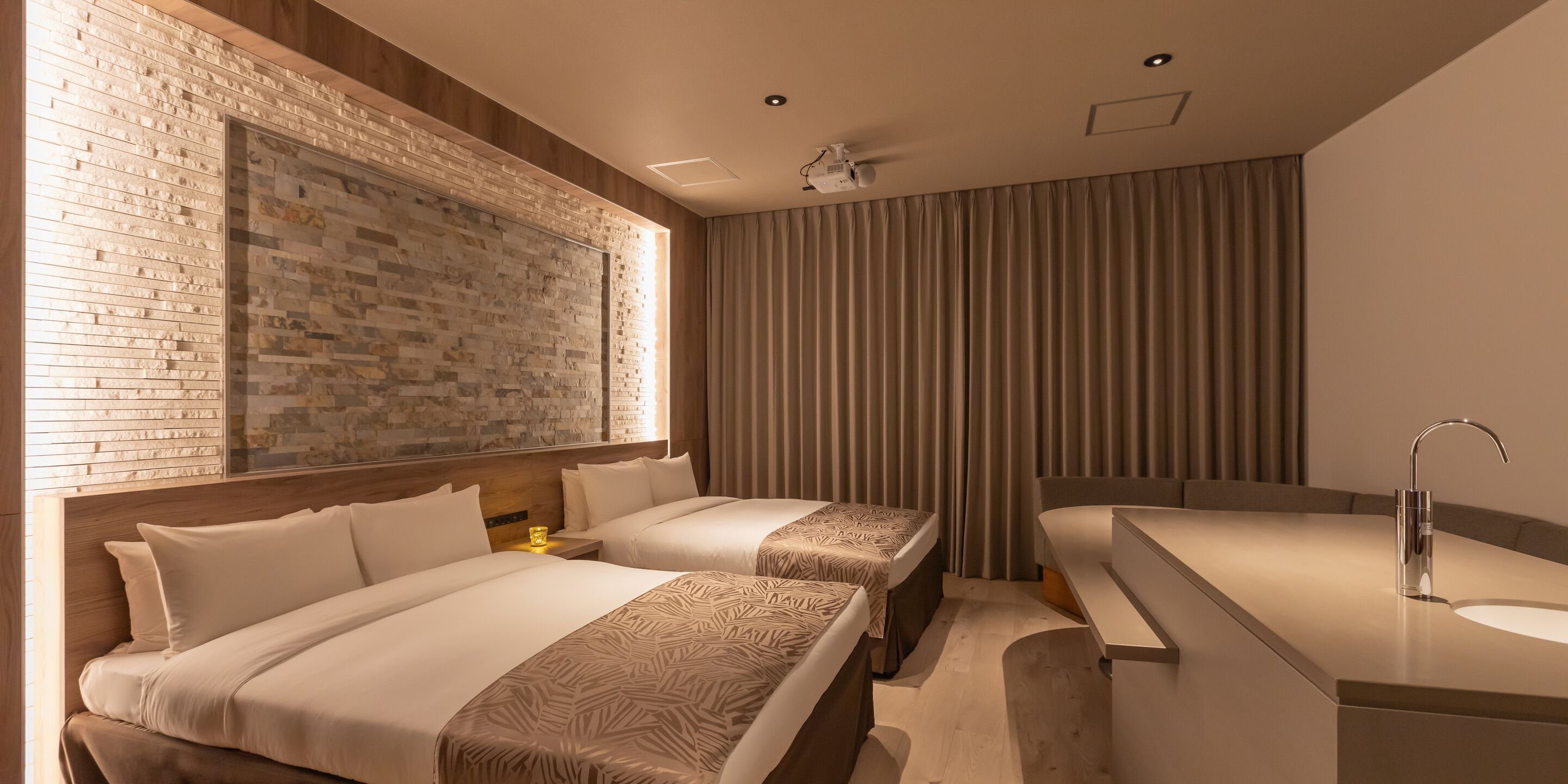 A hotel room with two beds, a large window with a brick wall, a modern bathroom with a sink and mirror, and a ceiling projector.