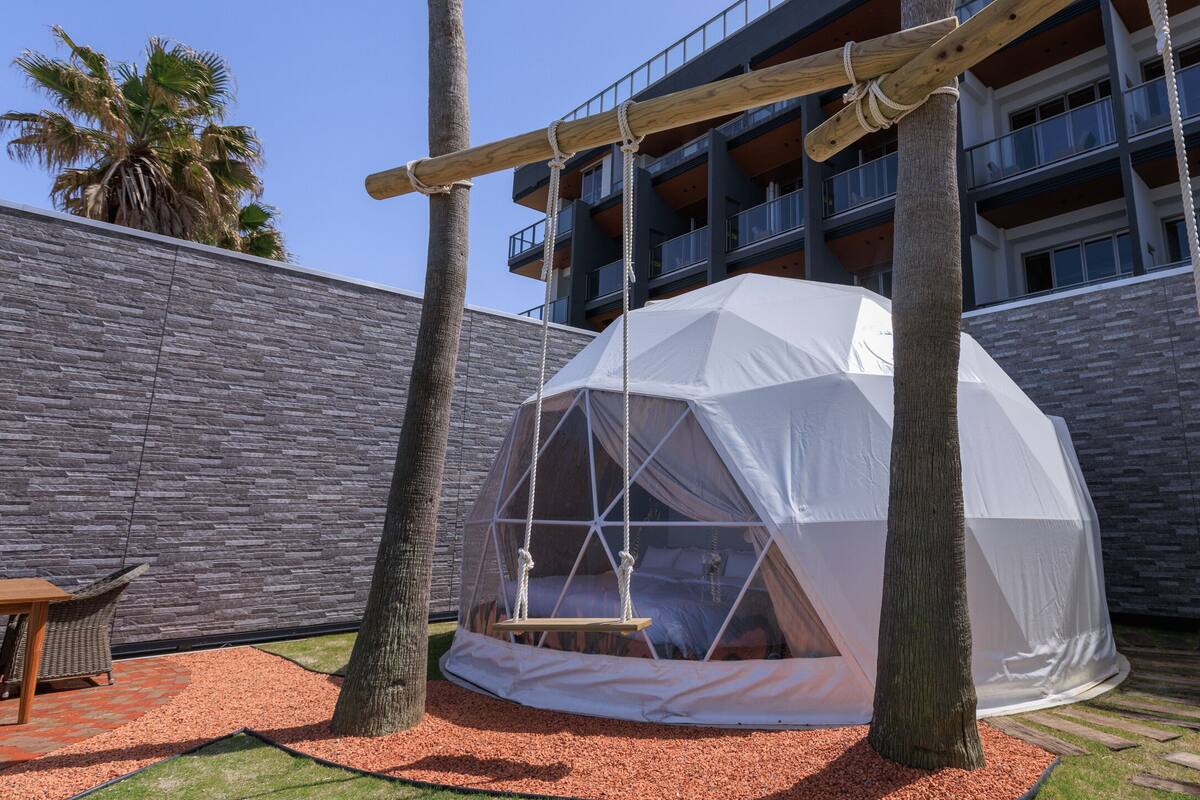 A white tent with a swing, a wooden table, and a stone wall in a courtyard.