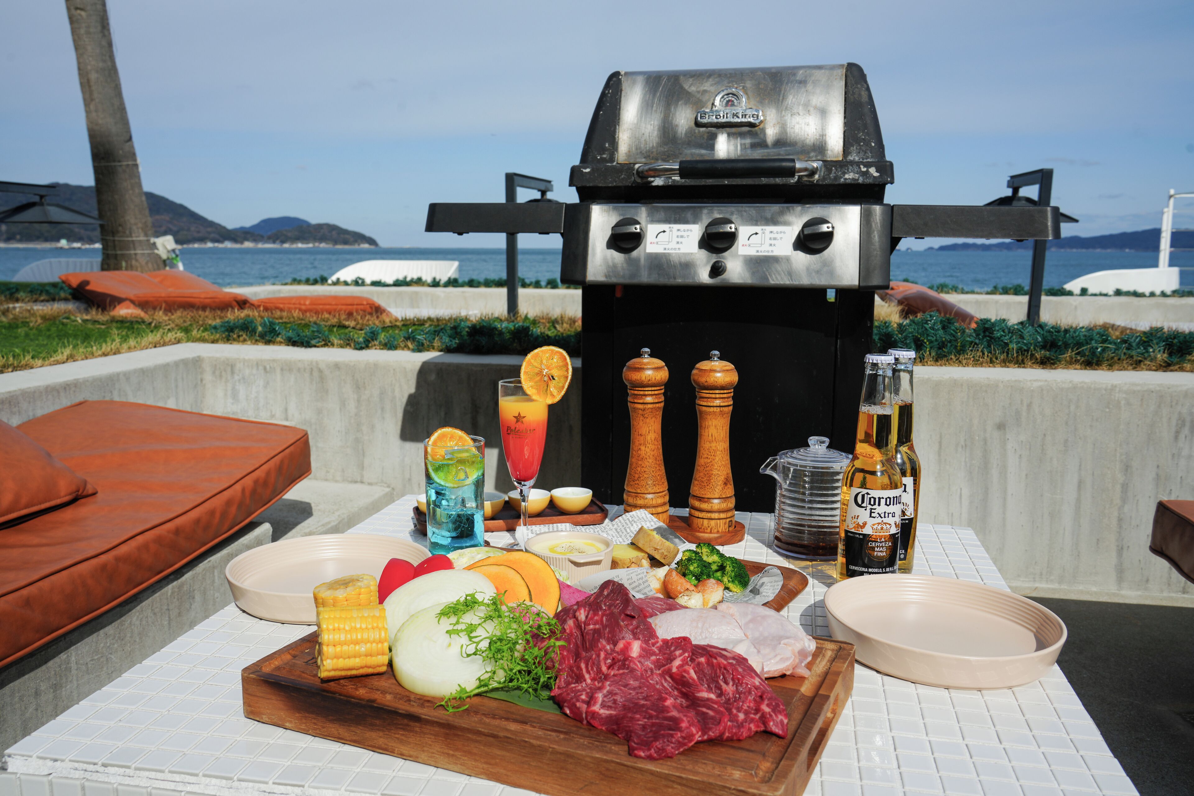 An outdoor dining area with a grill, various foods, drinks, and a view of the ocean.