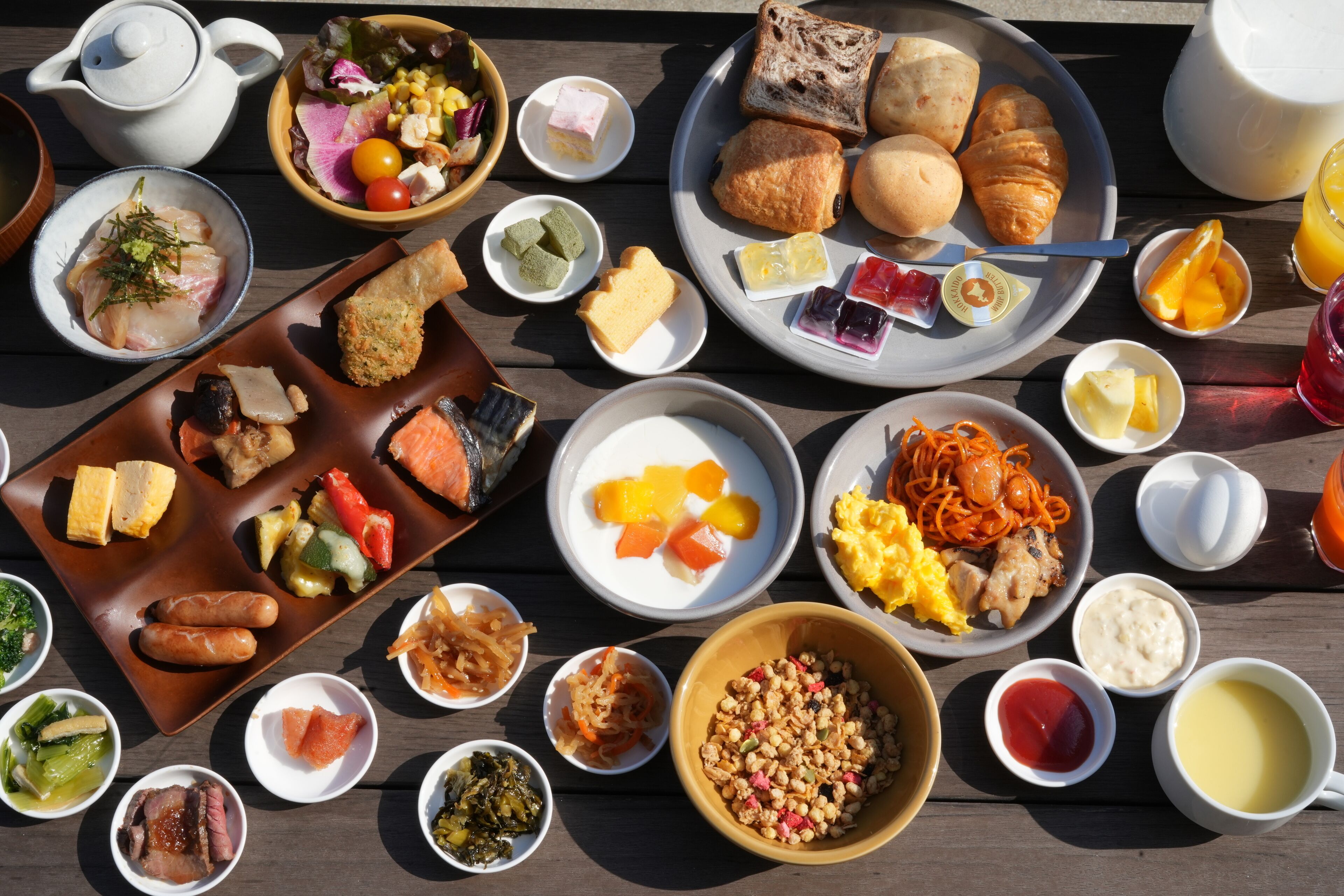 A variety of dishes and drinks on a wooden table, including bread, pastries, and a teapot.