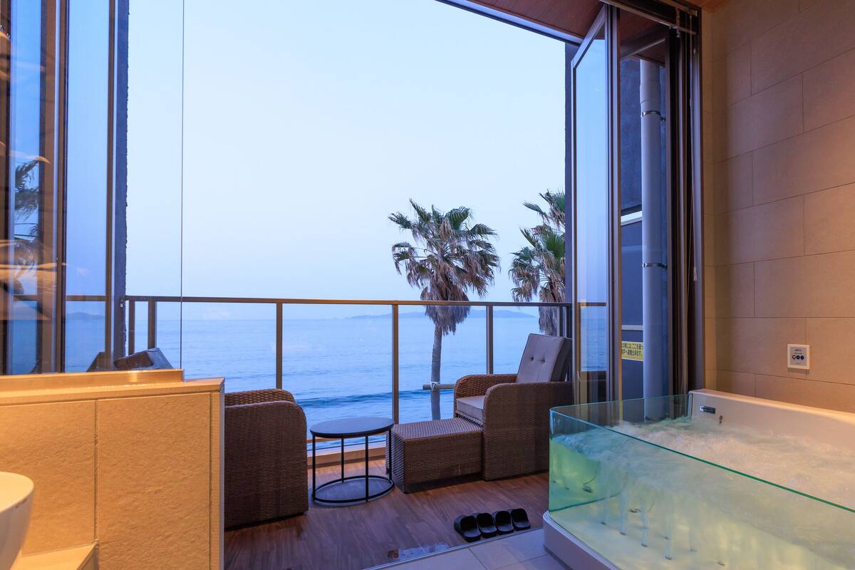 A balcony with a view of the ocean, a glass-enclosed bathtub, and wicker furniture.