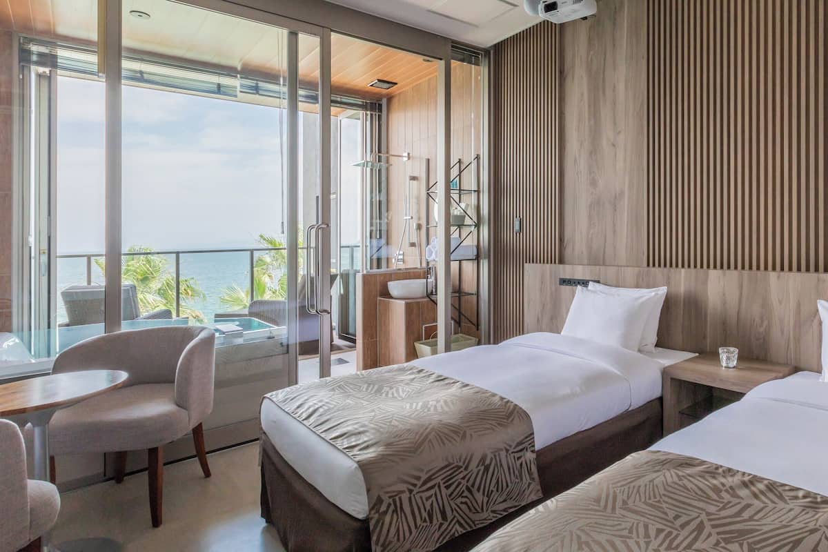 Habitación de hotel con dos camas, un balcón con vistas, una mesita pequeña y una silla.