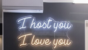 Un fauteuil noir et blanc à motifs, un pouf assorti et un panneau néon avec l’inscription « Je vous accueille, je vous aime », le tout placé devant un mur sombre.