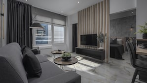 Een moderne woonkamer met een grijze bank, een flatscreen televisie, een eethoek met een tafel en stoelen, en een groot raam met gordijnen.