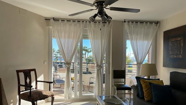 Un salon avec un ventilateur de plafond, un canapé, une table basse et un fauteuil. On peut voir un balcon aménagé avec du mobilier d’extérieur.