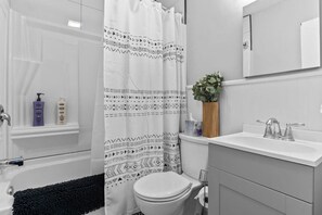 Une salle de bain moderne avec une baignoire, des toilettes et un lavabo. On y trouve un rideau de douche à motifs géométriques, une plante en pot et des articles de toilette sur le lavabo.