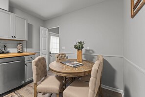 A dining area with a round wooden table, four upholstered chairs, a vase with greenery, and a kitchen in the background.