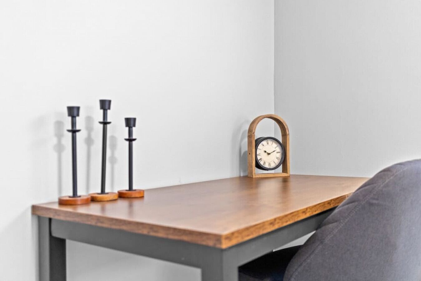 Een houten tafel met een klok en drie kandelaars voor een witte muur.