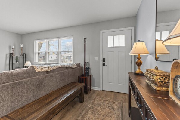A living room with a sofa, bench, wooden coffee table, and a lamp. There is a door with a mirror and a small table with a clock and decorative items. - Boho Chic Work Escape - 2-Bedroom, Close to Burlington/Airport (South Burlington)