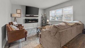A living room with a brown leather sofa, a brown armchair, a flat-screen TV mounted on the wall, a fireplace, and a large window with blinds. - Boho Chic Work Escape - 2-Bedroom, Close to Burlington/Airport (South Burlington)