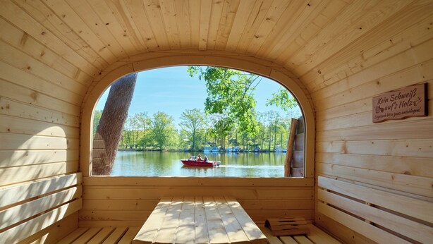 Une cabane en bois avec un banc et une table, et une grande fenêtre en arc qui donne sur un lac où se trouve un bateau.