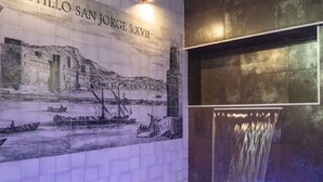 Eine mit Fliesen verkleidete Wand mit einer historischen Wandmalerei, die eine Küstenszene zeigt, ein gefliester Duschbereich und eine dunkle Wand mit Fliesen.