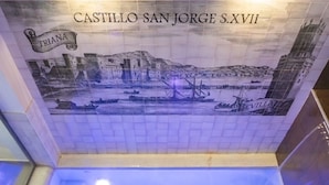 Eine mit Fliesen versehene Wand mit einem historischen Wandgemälde, das Castillo San Jorge XVI, Triana und Sevilla zeigt.