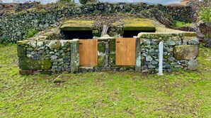 A stone structure with two wooden doors, surrounded by a grassy area and a stone wall. - Casa do Avô José Alves (T3), Country House - Rural Tourism  (Cabo da Praia)
