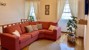 A cozy living room with a red sectional sofa, a wooden ceiling, and a large window with curtains. - Casa do Avô José Alves (T3), Country House - Rural Tourism  (Cabo da Praia)