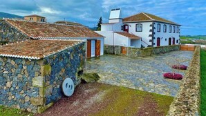 A stone building with a tiled roof, a white building with a red door, and a paved courtyard with a stone wall and a small garden. - Casa do Avô José Alves (T3), Country House - Rural Tourism  (Cabo da Praia)