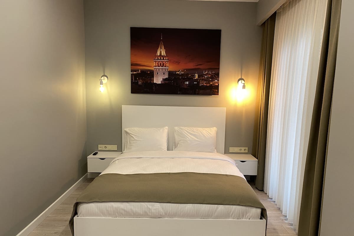 Una habitación de hotel moderna con una cama grande, dos mesitas de noche, un paragüero y un cuadro de paisaje urbano en la pared, con vistas a la ciudad a través de cortinas translúcidas.