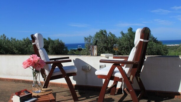 Zwei Holz-Loungesessel mit weißen Kissen, ein kleiner Tisch, eine Vase mit rosa Blumen und ein Buch auf dem Tisch. Im Hintergrund ein klarer, blauer Himmel und Blick aufs Meer.