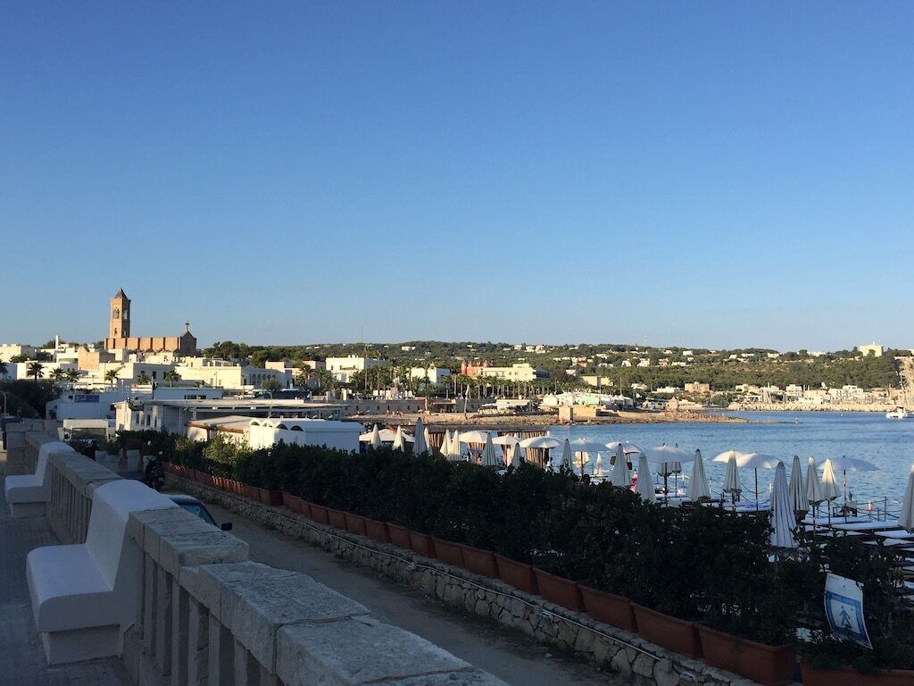A coastal town with a prominent church tower, a promenade with seating, and a marina with umbrellas.
