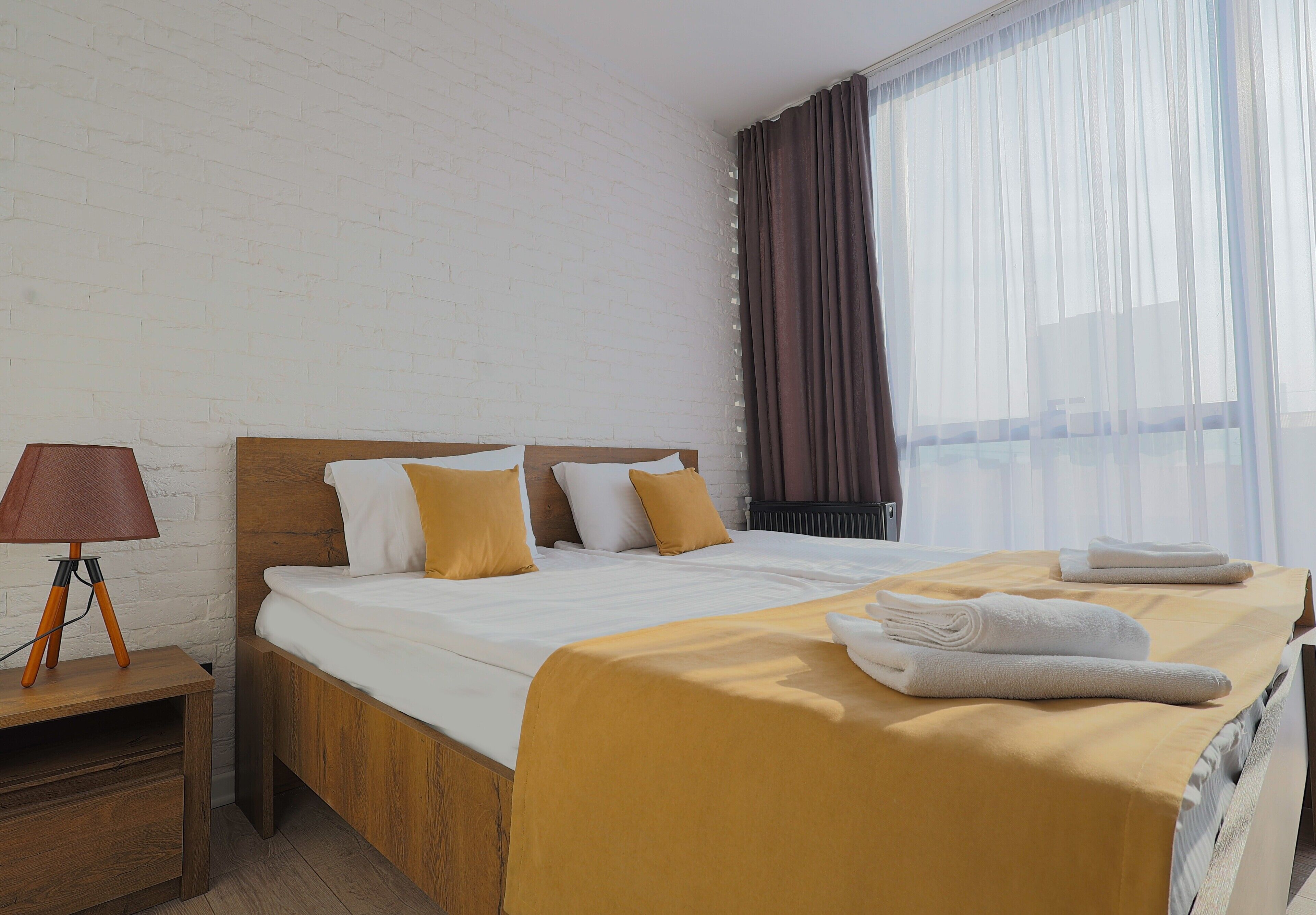 A hotel room with a wooden bed, white bedding, a wooden nightstand with a lamp, and a window with sheer curtains.