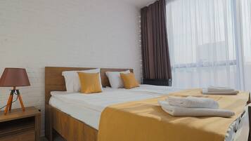 A hotel room with a wooden bed, white bedding, a wooden nightstand with a lamp, and a window with sheer curtains.
