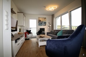 A modern living room with a sofa, a coffee table, and a blue armchair. There is a painting on the wall and a window with a view. - Sonnenweg 1 (Diemelsee)