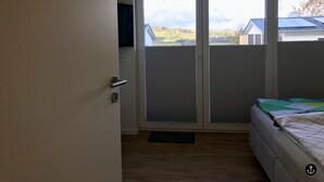 A bedroom with a sliding glass door leading to a balcony, a bed with a green and white quilt, and a wall-mounted TV. - Sonnenweg 1 (Diemelsee)