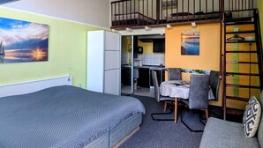 Ett kompakt rum med en säng, en soffa, ett matbord med stolar, en liten köksdel och en trappa upp till ett övre plan.