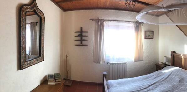 Een slaapkamer met een bed, een spiegel, een radiator, een raam met gordijnen en een klein nachtkastje.