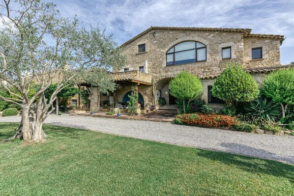 A stone building with a covered patio, a gravel driveway, and various plants and trees.