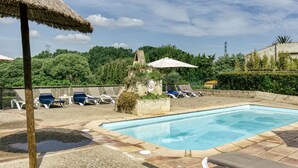 Piscina con sillones de descanso y sombrillas, rodeada de follaje y un muro de piedra.