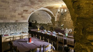 Un comedor de paredes de piedra con mesas dispuestas para una comida, sillas de madera y una araña.