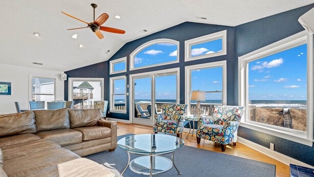 A spacious living room with a large glass table, comfortable seating, and a view of the ocean through large windows. - Sandbox 8 Bedroom Home in Kitty Hawk Shores (Kill Devil Hills)