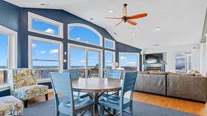 A spacious living area with a large window overlooking the ocean, a ceiling fan, and a modern kitchen. - Sandbox 8 Bedroom Home in Kitty Hawk Shores (Kill Devil Hills)
