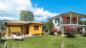 A two-story house with a covered patio, a lawn, and potted plants. - Holiday Home 'Garden House Lefkada' with Shared Terrace, Wi-Fi and Air Conditioning (Lefkada)