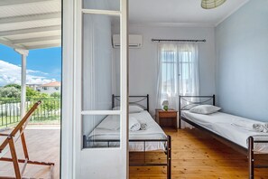 A room with two single beds, a wooden floor, a window with curtains, a small table with a lamp, and an air conditioning unit.