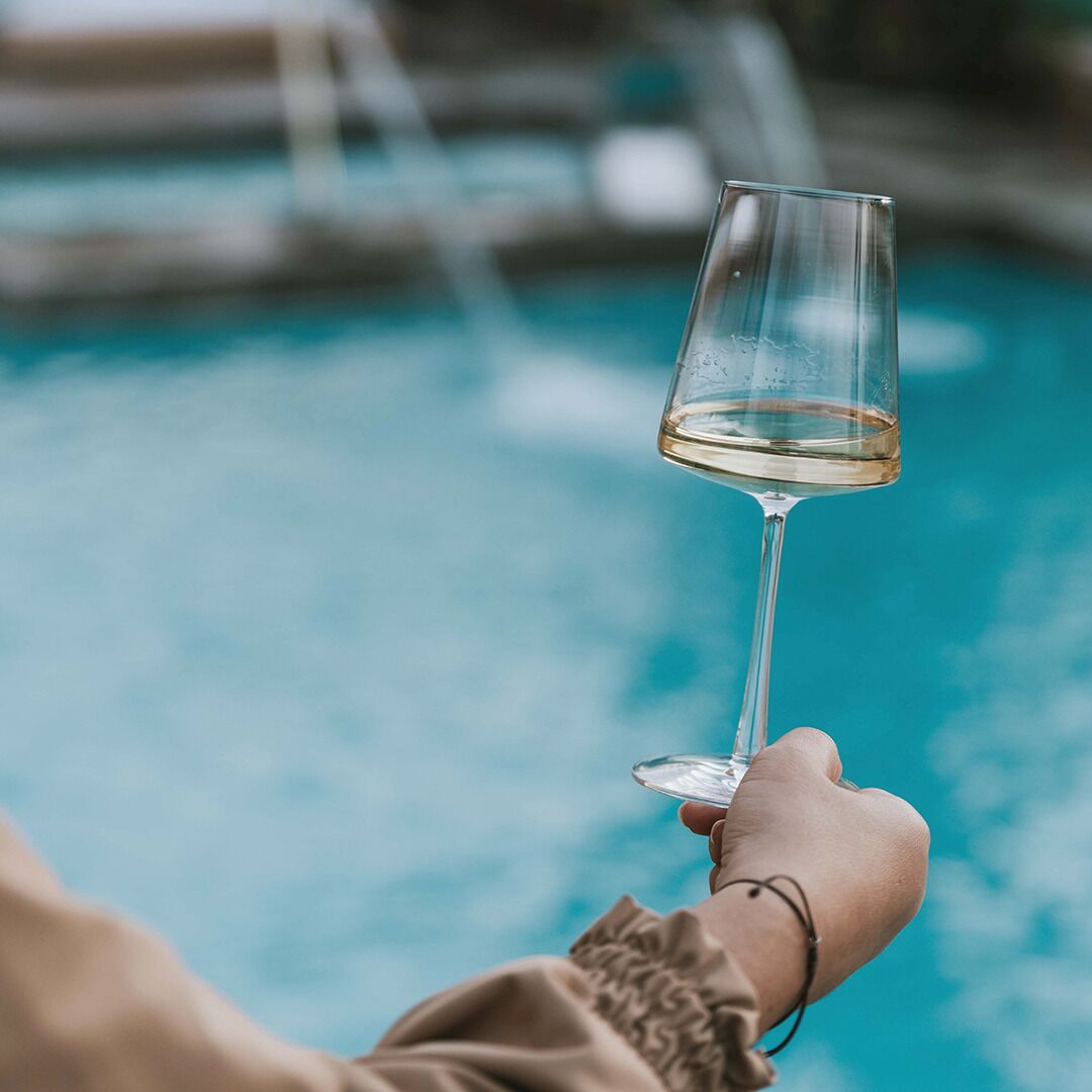 A hand holding a glass of white wine near a pool.