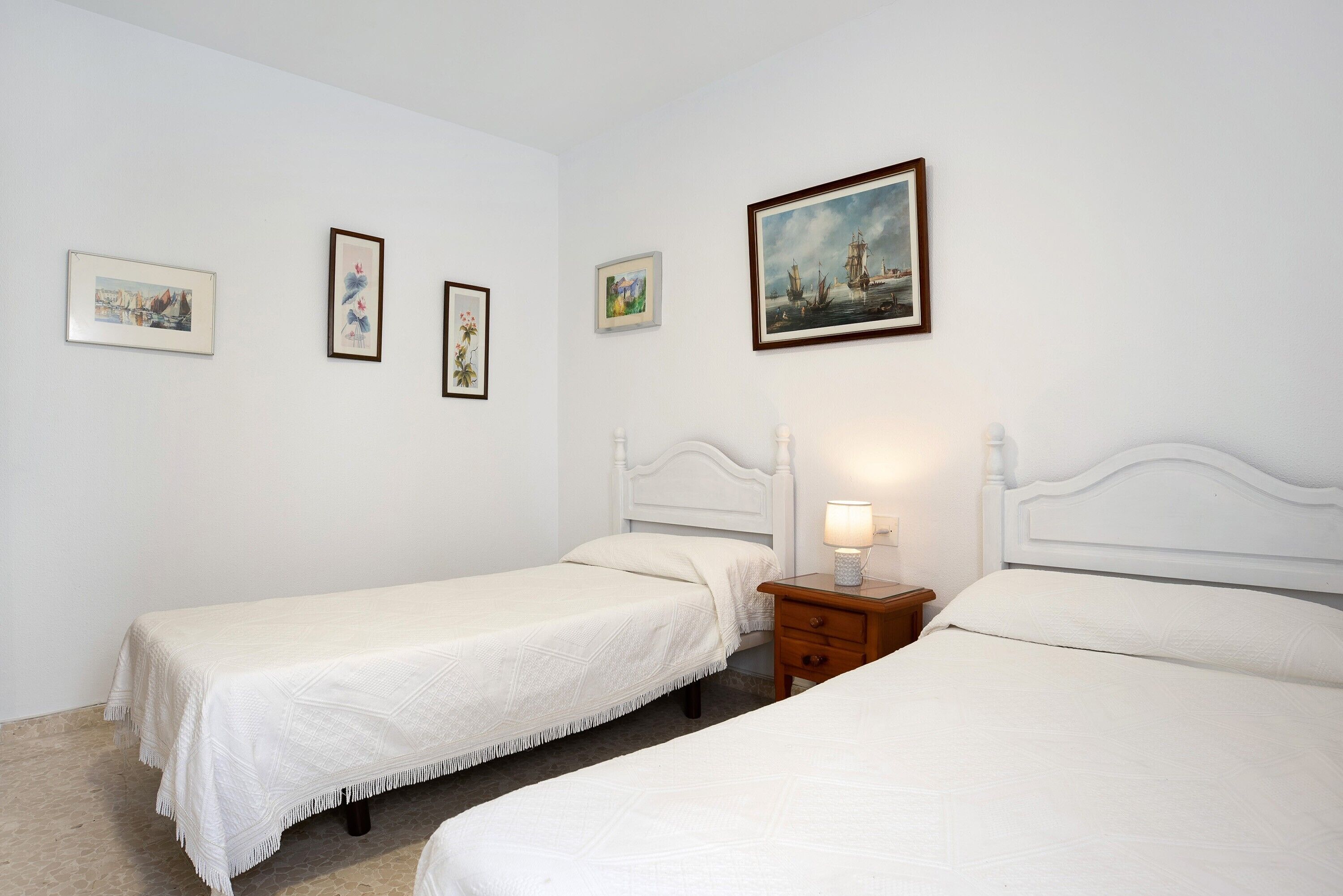 Une chambre avec deux lits simples, une table de chevet avec une lampe et trois tableaux encadrés accrochés au mur.
