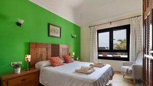 Una camera da letto con parete verde, testiera in legno, due cuscini, un letto con asciugamani, un comodino con lampada, una sedia e una finestra con tende.