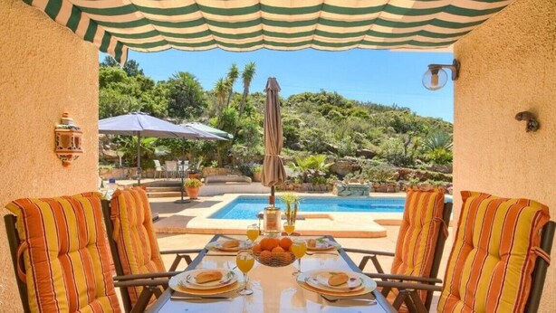 Eine Terrasse mit Tisch für zwei Personen, umgeben von gestreiften Stühlen, mit Pool und Grünanlagen im Hintergrund.