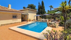 Ein Schwimmbecken mit Liegestühlen und Sonnenschirmen, umgeben von einem Garten mit Steinen und Pflanzen.