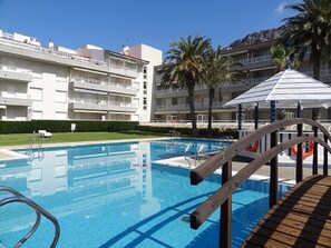 A swimming pool with a wooden deck, surrounded by palm trees and a multi-story building with balconies.