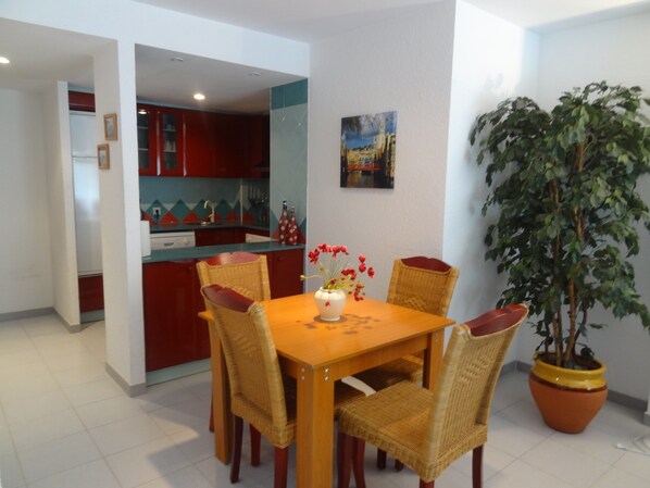 A dining area with a wooden table and wicker chairs, a potted plant, and a kitchen in the background.