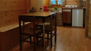 A wooden cabin interior with a dining table, chairs, a kitchenette, and a window with curtains.
