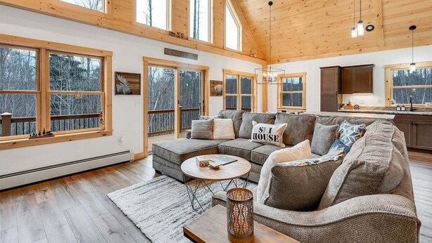 A spacious living room with a large sectional sofa, a round coffee table, and a fireplace. There are large windows offering views of the surrounding forest.