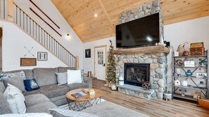 A living room with a stone fireplace, a large flat-screen TV, a wooden staircase, and a Christmas tree.