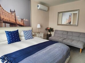 Habitación de hotel con cama, sofá gris, espejo, mesita de noche con lámpara y aire acondicionado instalado en la pared.