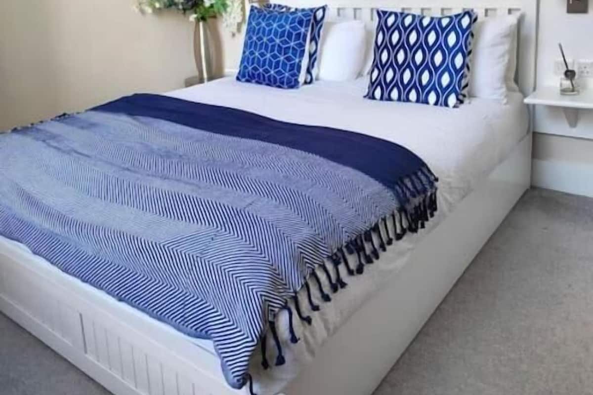 Una cama bien tendida con una colcha y almohadas estampadas, una mesita de noche con una lámpara y un reloj de pared.